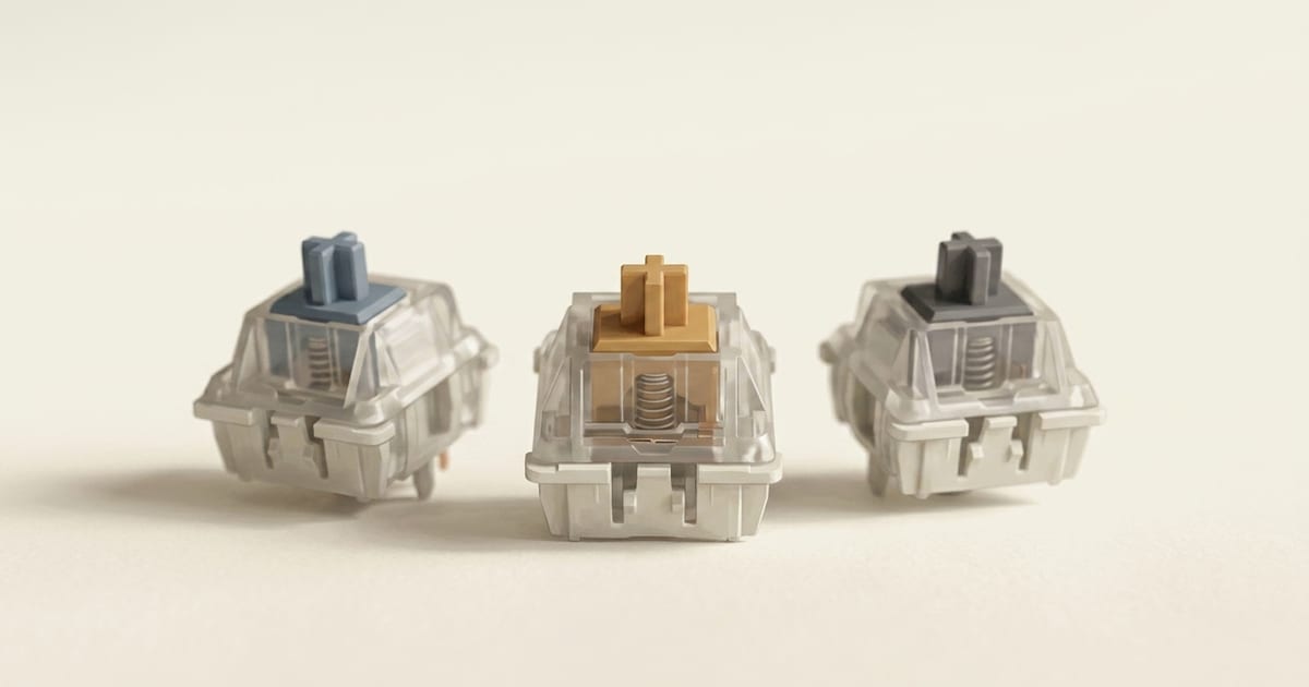 Three mechanical keyboard switches with translucent housings revealing internal springs, stems in amber, slate-blue, and charcoal.